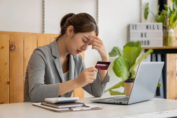 Young Asian woman having trouble paying internet shoppers Depressed woman sitting at desk with laptop holding credit card feeling depressed and worried because overspending on card