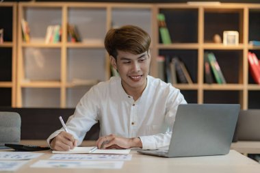 Serious young asian man working in office at desk. Holding and looking at document. Graph. Planning in office. calculation report Using laptop and calculator, online meeting concept