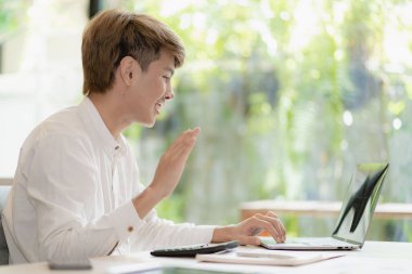 Serious young asian man working in office at desk. Holding and looking at document. Graph. Planning in office. calculation report Using laptop and calculator, online meeting concept