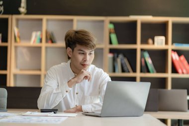 Serious young asian man working in office at desk. Holding and looking at document. Graph. Planning in office. calculation report Using laptop and calculator, online meeting concept