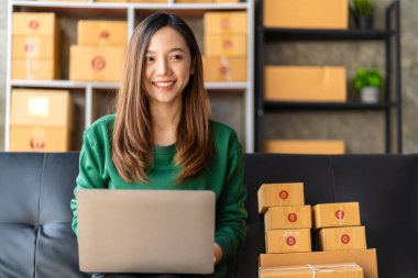 Küçük bir iş kurun, genç Asyalı bir kadın çevrimiçi alışveriş yaparak kutuları paketliyor ve dijital dizüstü bilgisayardan müşterilere teslim edilecek ürünleri kontrol ediyor..