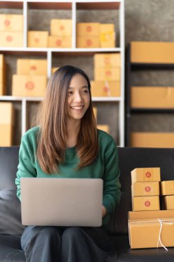 Küçük bir iş kurun, genç Asyalı bir kadın çevrimiçi alışveriş yaparak kutuları paketliyor ve dijital dizüstü bilgisayardan müşterilere teslim edilecek ürünleri kontrol ediyor..