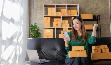 Heyecanlı Asyalı bayan küçük işletme sahibi akıllı telefon sahibi müşteriden mutlu bir şekilde isim siparişi alıyor. Ev alışverişinde dizüstü bilgisayarla çalışırken paket göndermeye hazırlanın.