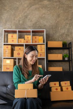 Küçük işletme sahibi konsepti. Akıllı telefonlu çekici Asyalı kadın mesajları mutlu bir şekilde kontrol ediyor. Evdeki online alışverişte dizüstü bilgisayarla çalışmak paketleri dikey fotoğraf dağıtmaya hazırlamak