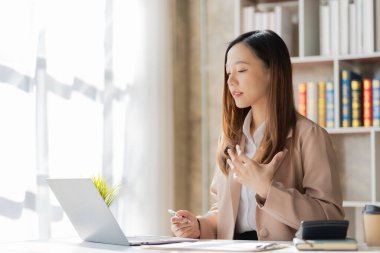 Neşeli Asyalı iş kadını dizüstü bilgisayarı ve finansal grafik kağıdıyla masada oturuyor. Finansal iş planı ve ofisteki online toplantıda kameraya gülümsüyor.