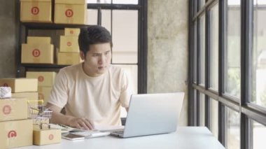 Tişörtlü genç Asyalı işadamı ev ofisinde dizüstü bilgisayarla çalışıyor, siparişleri alıyor, ismi paket kutusu teslimatı için iş yapıyor gibi görünüyor, KOBİ çevrimiçi pazarlama.