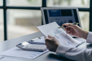 Asyalı iş kadınlarının ellerinin kalem tutması ve ofisindeki yatırım analizleri için finansal belgelere, yatırım planlarına ve grafik çizelgelerine işaret etmesi.