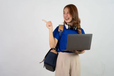 Attractive young Asian college student with a backpack on her shoulder making a gesture with pointing hand and holding a laptop in her hand. isolated on white background