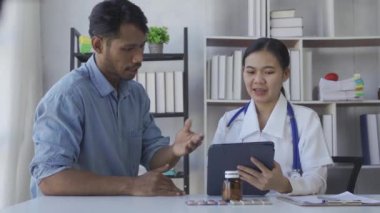 Beyaz önlüklü Asyalı kadın doktor, pano ve tablet kullanarak sağlık ocağındaki masada oturan erkek hastayla sonuçları veya belirtileri tartışıyor.