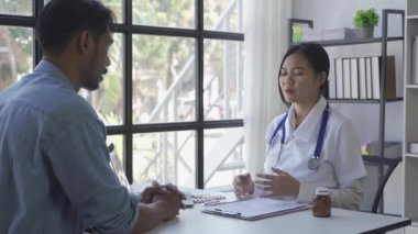 Asian female doctor in white medical coat using clipboard and tablet discussing results or symptoms with male patient sitting at table in health clinic