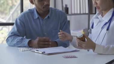 Beyaz önlüklü Asyalı kadın doktor, pano ve tablet kullanarak sağlık ocağındaki masada oturan erkek hastayla sonuçları veya belirtileri tartışıyor.