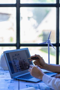 Male engineer meeting pointing at blueprint with house plan and wind turbine next to house building concept with wind turbine renewable energy installation