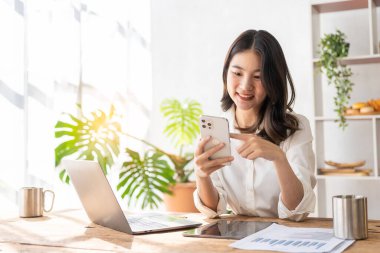 Beautiful Asian female accountant working with laptop at her desk analyzing business reports with folders, files and documents. Mobile smartphone messaging or text chatting. online work concept