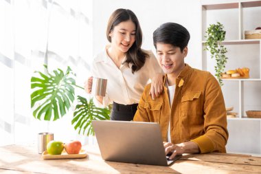 Cheerful young Asian couple working together in cozy living room at home. Couple working at home with laptop, financial accounting concept, pie in house