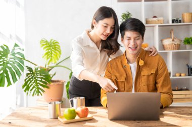 Cheerful young Asian couple working together in cozy living room at home. Couple working at home with laptop, financial accounting concept, pie in house