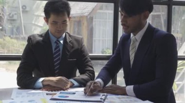 Two businessmen in suits discussing project on paper with graphs and charts. Pointing to Financial Documents in Dark Office 4K