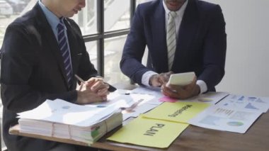 Two businessmen in suits discussing project on paper with graphs and charts. Pointing to Financial Documents in Dark Office 4K