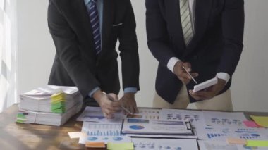 Two businessmen in suits discussing project on paper with graphs and charts. Pointing to Financial Documents in Dark Office 4K