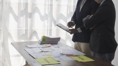 Two businessmen in suits discussing project on paper with graphs and charts. Pointing to Financial Documents in Dark Office 4K