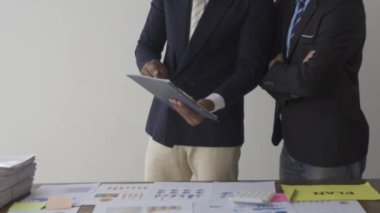 Two businessmen in suits discussing project on paper with graphs and charts. Pointing to Financial Documents in Dark Office 4K