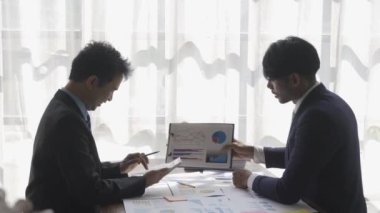 Two businessmen in suits discussing project on paper with graphs and charts. Pointing to Financial Documents in Dark Office 4K