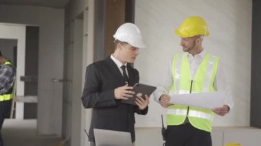 Engineer and businessman in hard hat meeting and using computer tablet and print documents in unfinished buildings under construction on the background, in 4K (UHD).