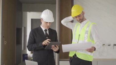 Engineer and businessman in hard hat meeting and using computer tablet and print documents in unfinished buildings under construction on the background, in 4K (UHD).