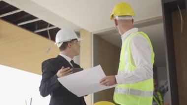 Engineer and businessman in hard hat meeting and using computer tablet and print documents in unfinished buildings under construction on the background, in 4K (UHD).
