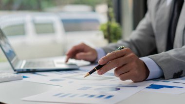 Businessman hand holding a pen pointing in document, financial graph, plan, marketing graph. Financial investment and business strategy data chart report and working on laptop in office