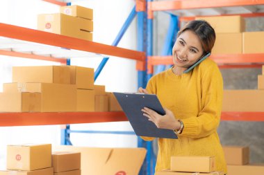 Asian woman holding smartphone talking to customer, checking delivery box, online sale, parcel delivery business concept, SME business startup