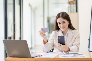 Masasında oturan Asyalı iş kadını internet alışverişini ödemek için mobil bankacılık kullanıyor. Laptop ve finansal belgelerle birlikte çalışma masasında.