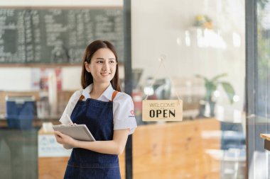 Beautiful Asian barista in apron holding tablet and standing in front of cafe door with open sign Concept of food and beverage business