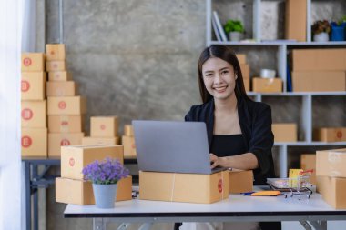 Evde dizüstü bilgisayar ve kutularla çalışan genç Asyalı kadın siparişleri kontrol ediyor ve paket teslimatı hazırlıyor, KOBİ çevrimiçi iş kavramı
