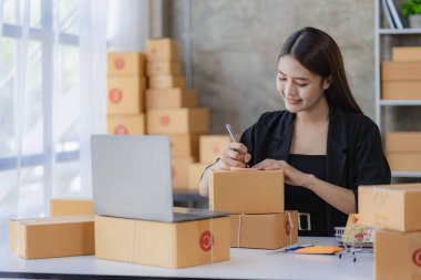 Evde dizüstü bilgisayar ve kutularla çalışan genç Asyalı kadın siparişleri kontrol ediyor ve paket teslimatı hazırlıyor, KOBİ çevrimiçi iş kavramı
