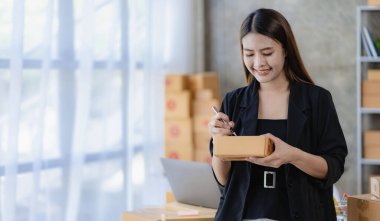 Evde dizüstü bilgisayar ve kutularla çalışan genç Asyalı kadın siparişleri kontrol ediyor ve paket teslimatı hazırlıyor, KOBİ çevrimiçi iş kavramı