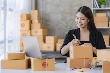 Evde dizüstü bilgisayar ve kutularla çalışan genç Asyalı kadın siparişleri kontrol ediyor ve paket teslimatı hazırlıyor, KOBİ çevrimiçi iş kavramı