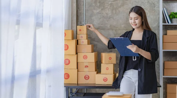 Young Asian woman working from home with laptop and boxes checking orders and preparing parcel delivery, SME online business concept