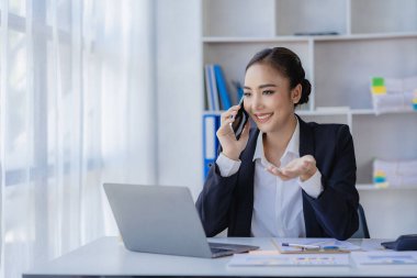 Serbest çalışan Asyalı iş kadını telefonla konuşuyor ve dizüstü bilgisayarla ve finansal grafik belgeleriyle çalışıyor. İşyerindeki işlerini yönetiyor. Ofisteki mali raporlarla ilgili bir plan yapıyor..
