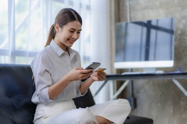 Kanepede oturan Asyalı kadın internete akıllı telefon ve kredi kartıyla ödeme yapıyor, online alışveriş kavramı.