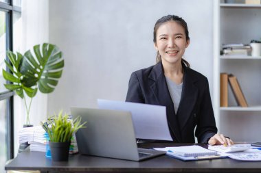 Gülümseyen Asyalı iş kadını, modern ofis ofisindeki bilgisayarında çalışıyor. Hesaplamalar yapıyor. Masrafları analiz ediyor.