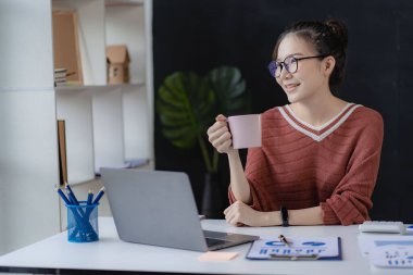 Gülümseyen Asyalı iş kadını, modern ofis ofisindeki bilgisayarında çalışıyor. Hesaplamalar yapıyor. Masrafları analiz ediyor.