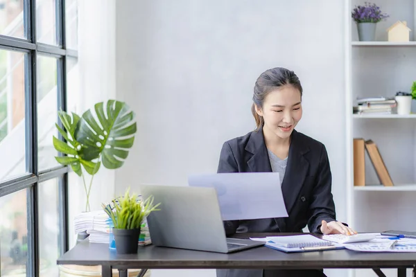 stock image Smiling Asian businesswoman working on laptop computer at her desk in modern home office doing calculations analyzing expenses reporting finances