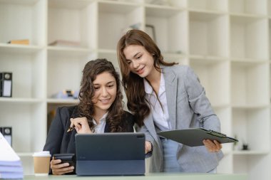 Konferans masasında dijital tablet kullanarak çalışan iki neşeli iş kadını ve emlak projesi görüşmesi için ofis iş arkadaşlarıyla birlikte çalışan belgeler.