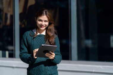 Ofisin önünde elektronik tablet tutan uzun saçlı güzel bir kadın. Uzun kahverengi saçlı başarılı bir iş kadını. Bina yakınında ve elinde tabletle çalışıyor.