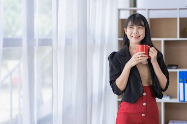 Milenyum başarılı Asyalı kadın sekreter resmi takım elbiseli kurumsal ofiste duran bir fincan kahveyi tutuyor.