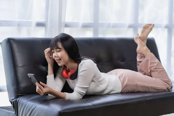 stock image Happy and beautiful Asian woman wearing headphones Select a music playlist on your smartphone. Listening to music while lying on the sofa in the living room at home, relaxation concept.