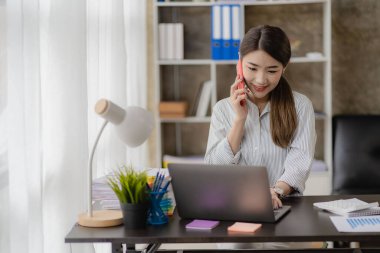 Asyalı iş kadını, evden akıllı bir şekilde çalışırken iş arkadaşlarıyla görüntülü görüşme planları hakkında konuşmak için internet üzerinden akıllı telefon kullanıyor..