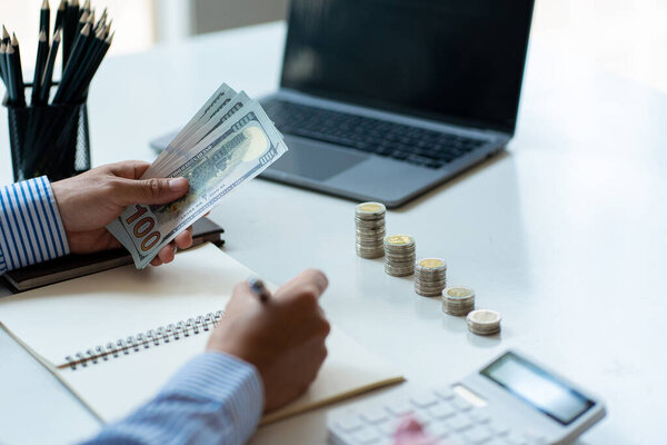 Young man is using pen to record amount of money and coins for accounting plan on wooden table in home office, accounting and finance calculation concept