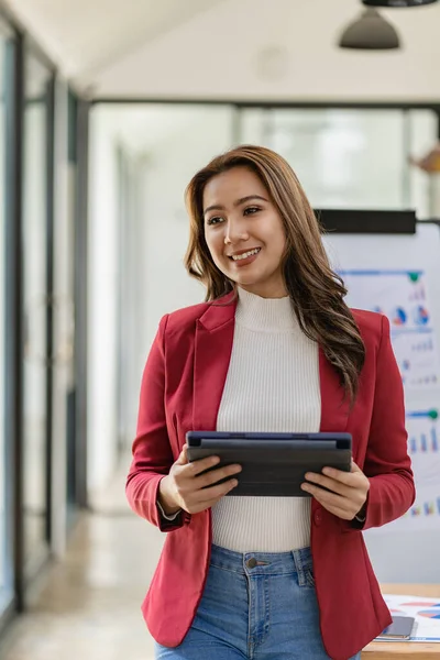 stock image Pretty and attractive asian businesswoman or female clerk in formal suit using digital tablet standing in front of her desk, financial accounting concept