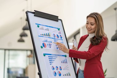 Takım elbiseli Asyalı iş kadını elinde kalemle konferans odasında verileri sunan beyaz tahtadaki belge grafiğine bakıyor.